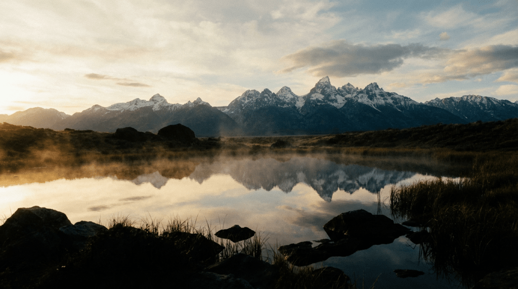 Reflection of snow-covered mountain peaks in a still pond with rising morning mist.
